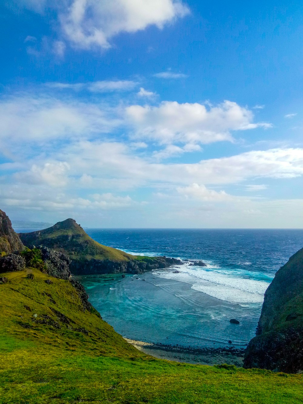 green grass field on mountain near body of water under blue and white cloudy sky during