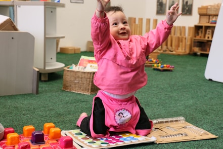 A young child wearing a bright pink outfit is sitting on the floor in a playful pose, with arms raised and a joyful expression. Surrounding the child are colorful toys and educational materials. The setting appears to be a classroom or playroom with toys, books, and wooden shelving in the background.