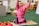 A young child wearing a bright pink outfit is sitting on the floor in a playful pose, with arms raised and a joyful expression. Surrounding the child are colorful toys and educational materials. The setting appears to be a classroom or playroom with toys, books, and wooden shelving in the background.
