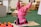 A young child wearing a bright pink outfit is sitting on the floor in a playful pose, with arms raised and a joyful expression. Surrounding the child are colorful toys and educational materials. The setting appears to be a classroom or playroom with toys, books, and wooden shelving in the background.
