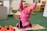 A young child wearing a bright pink outfit is sitting on the floor in a playful pose, with arms raised and a joyful expression. Surrounding the child are colorful toys and educational materials. The setting appears to be a classroom or playroom with toys, books, and wooden shelving in the background.