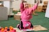 A young child wearing a bright pink outfit is sitting on the floor in a playful pose, with arms raised and a joyful expression. Surrounding the child are colorful toys and educational materials. The setting appears to be a classroom or playroom with toys, books, and wooden shelving in the background.