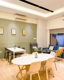 A modern interior space featuring two tables with chairs, a blue couch with pillows, and a wooden rocking chair. Green plants in vases decorate the tables. The walls are painted in light gray with framed artworks. The room has wooden flooring and is lit by ceiling lights.