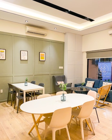 A modern interior space featuring two tables with chairs, a blue couch with pillows, and a wooden rocking chair. Green plants in vases decorate the tables. The walls are painted in light gray with framed artworks. The room has wooden flooring and is lit by ceiling lights.
