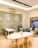 A modern interior space featuring two tables with chairs, a blue couch with pillows, and a wooden rocking chair. Green plants in vases decorate the tables. The walls are painted in light gray with framed artworks. The room has wooden flooring and is lit by ceiling lights.