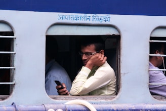 A man is sitting inside a train, holding a phone to his ear, possibly engaged in a call. The window has bars, and there are two other passengers partially visible, also appearing to be using their phones or listening to something. The train has text written above the window in a non-English script, possibly indicating the destination or service.