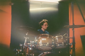 A person wearing glasses is playing a drum set in a dimly lit room. The drums are in the foreground, and the background features contrasting dark and orange colors.