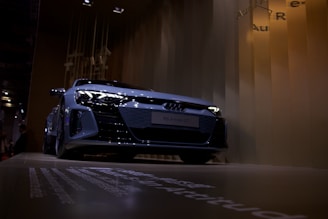 A sleek silver car is displayed against a modern, angular background with soft lighting. The low-angle shot emphasizes the car's aerodynamic design and highlights its glossy surface.
