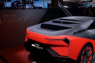 A futuristic concept car with sleek, angular design and vibrant red accents is displayed indoors. The vehicle features a dark gray body with a stylish rear and innovative detailing, adding to its modern look. The raised gullwing door and illuminated branding emphasize its advanced technology.