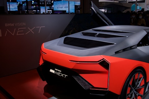 A futuristic concept car with sleek, angular design and vibrant red accents is displayed indoors. The vehicle features a dark gray body with a stylish rear and innovative detailing, adding to its modern look. The raised gullwing door and illuminated branding emphasize its advanced technology.