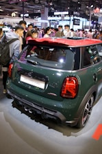 A John Cooper Works car is displayed in a showroom setting, surrounded by a crowd of people who appear to be interested in the vehicle. The car has a dark green body with a contrasting red roof, and is positioned under bright lighting. The setting suggests an automotive event or exhibition with various other displays visible in the background.