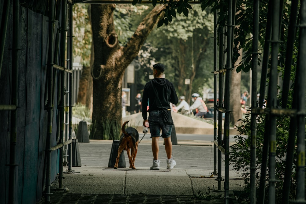 man in black jacket and blue denim jeans walking with brown short coated dog on sidewalk