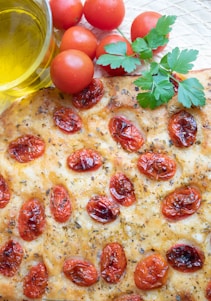 A close-up view of a freshly baked focaccia bread topped with cherry tomatoes, herbs, and a drizzle of olive oil. The bread is golden brown with a crispy texture, and the tomatoes are roasted to a vibrant red. Nearby, there are fresh whole tomatoes and sprigs of parsley, accompanied by a glass dish containing olive oil.
