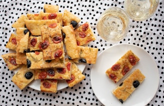A collection of rectangular focaccia pieces topped with cherry tomatoes and olives is arranged on a white plate. Beside it, two glasses of white wine are placed on a dotted fabric background. Another smaller white plate holds two additional pieces of focaccia.