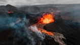 A dramatic volcanic eruption emits bright orange lava spewing from the crater, with rivers of molten rock flowing down the dark, rocky landscape. Dense smoke and ash billow into the sky, partially obscuring the rugged terrain and distant mountains under overcast clouds.