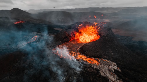 A dramatic volcanic eruption emits bright orange lava spewing from the crater, with rivers of molten rock flowing down the dark, rocky landscape. Dense smoke and ash billow into the sky, partially obscuring the rugged terrain and distant mountains under overcast clouds.
