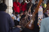 A group of men is gathered in a semi-circle, with one prominently holding a traditional stringed musical instrument. The setting appears to be indoors, possibly in a cultural or religious building, featuring decorative elements. The focus is on the musician, while others around him seem attentive or deeply engaged. The clothing is varied, with some wearing bright red, adding a splash of color.