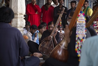 A group of men is gathered in a semi-circle, with one prominently holding a traditional stringed musical instrument. The setting appears to be indoors, possibly in a cultural or religious building, featuring decorative elements. The focus is on the musician, while others around him seem attentive or deeply engaged. The clothing is varied, with some wearing bright red, adding a splash of color.
