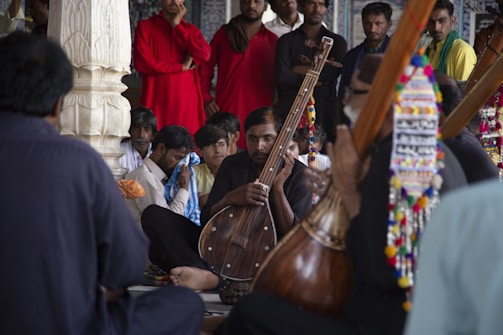 A group of men is gathered in a semi-circle, with one prominently holding a traditional stringed musical instrument. The setting appears to be indoors, possibly in a cultural or religious building, featuring decorative elements. The focus is on the musician, while others around him seem attentive or deeply engaged. The clothing is varied, with some wearing bright red, adding a splash of color.