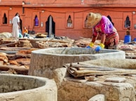 A person wearing a straw hat and brightly colored clothes is standing by a stone vat, engaging in work that appears to involve processing materials, possibly leather. Several large stone vats and piles of leather or fabric are spread across an outdoor workspace. The background features a terracotta building with pointed arched windows, enhancing the traditional atmosphere. Additional workers are visible further back, contributing to an active scene.