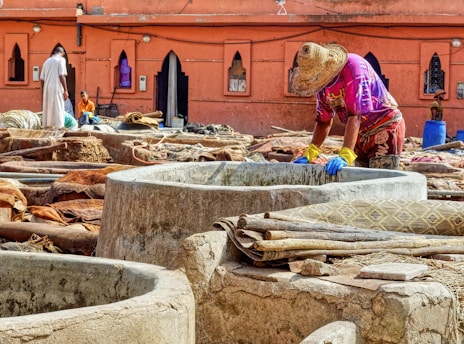 A person wearing a straw hat and brightly colored clothes is standing by a stone vat, engaging in work that appears to involve processing materials, possibly leather. Several large stone vats and piles of leather or fabric are spread across an outdoor workspace. The background features a terracotta building with pointed arched windows, enhancing the traditional atmosphere. Additional workers are visible further back, contributing to an active scene.