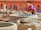 A person wearing a straw hat and brightly colored clothes is standing by a stone vat, engaging in work that appears to involve processing materials, possibly leather. Several large stone vats and piles of leather or fabric are spread across an outdoor workspace. The background features a terracotta building with pointed arched windows, enhancing the traditional atmosphere. Additional workers are visible further back, contributing to an active scene.