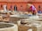 A person wearing a straw hat and brightly colored clothes is standing by a stone vat, engaging in work that appears to involve processing materials, possibly leather. Several large stone vats and piles of leather or fabric are spread across an outdoor workspace. The background features a terracotta building with pointed arched windows, enhancing the traditional atmosphere. Additional workers are visible further back, contributing to an active scene.
