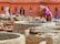 A person wearing a straw hat and brightly colored clothes is standing by a stone vat, engaging in work that appears to involve processing materials, possibly leather. Several large stone vats and piles of leather or fabric are spread across an outdoor workspace. The background features a terracotta building with pointed arched windows, enhancing the traditional atmosphere. Additional workers are visible further back, contributing to an active scene.