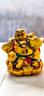 A golden laughing Buddha statue adorned with a black beaded necklace and holding a large gold ingot in one hand. The figure is seated on a pile of gold coins and wears a robe with red and green accents.