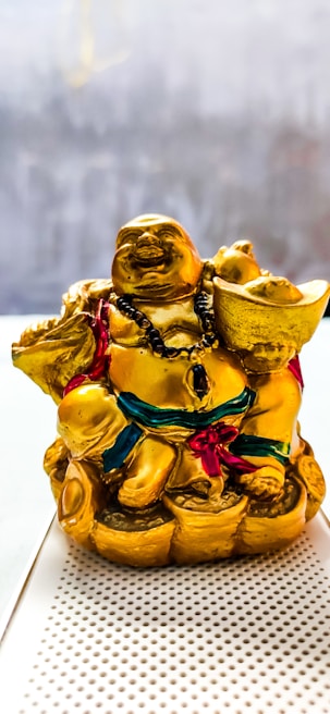 A golden laughing Buddha statue adorned with a black beaded necklace and holding a large gold ingot in one hand. The figure is seated on a pile of gold coins and wears a robe with red and green accents.
