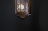A vintage wall clock with intricate gold patterns is mounted on a dark wall. The clock's wooden frame has an elegant, curved design at the base.