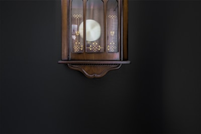 A vintage wall clock with intricate gold patterns is mounted on a dark wall. The clock's wooden frame has an elegant, curved design at the base.