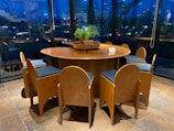 A round wooden table is surrounded by six wooden chairs with cushioned seats. The setting is indoors with large, clear glass windows that reveal a view of exterior lights and plants. On the center of the table is a planter with green foliage, accompanied by a small card displaying information.