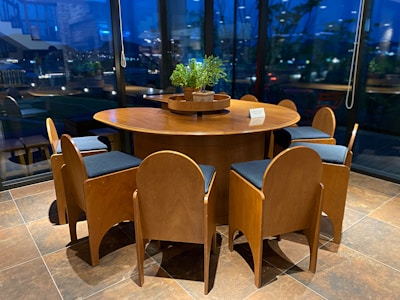 A round wooden table is surrounded by six wooden chairs with cushioned seats. The setting is indoors with large, clear glass windows that reveal a view of exterior lights and plants. On the center of the table is a planter with green foliage, accompanied by a small card displaying information.