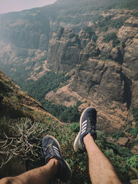 person wearing black and white adidas sneakers sitting on cliff