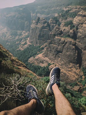 person wearing black and white adidas sneakers sitting on cliff