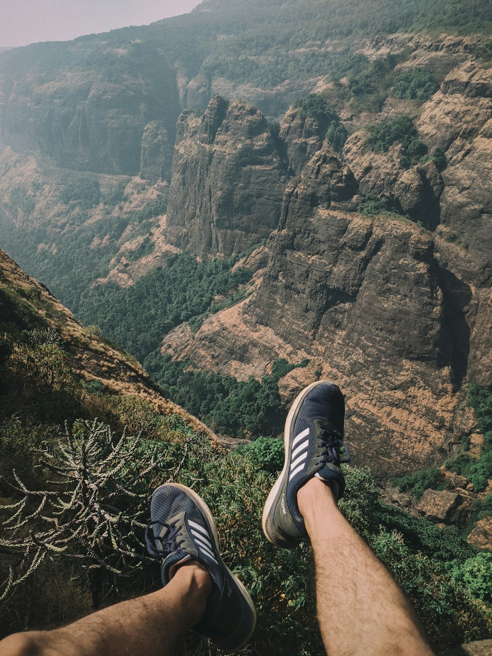 person wearing black and white adidas sneakers sitting on cliff
