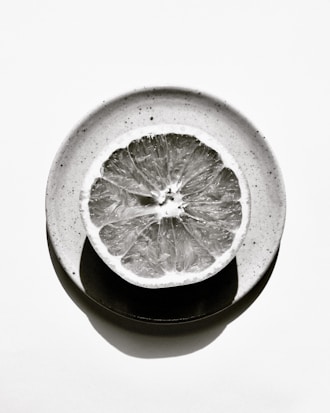 A monochrome image of a citrus fruit slice placed on a ceramic plate. The fruit's texture and intricate details are highlighted, with light and shadow creating a dramatic effect.