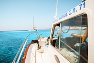 A close-up view of a boat with Greek writing on the roof in clear blue water. Another sailboat is anchored in the distance with mountains in the background under a bright, clear sky.
