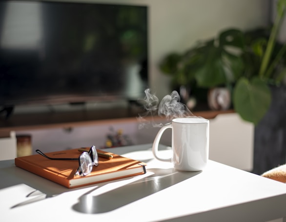 A cozy indoor setting featuring a white mug with steam rising from it, placed on a white table. A closed orange notebook with a pair of glasses rests nearby. In the background, there is a blurred television screen and lush green houseplants, creating a relaxing atmosphere filled with natural light.