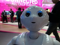 A humanoid robot with large, expressive eyes stands prominently, capturing attention in a tech event setting. People in business attire are visible in the background, engaged in conversation and examining displays. The scene is vibrant with a backdrop that features the phrase 'Connected All Over Europe' in bold pink lettering.