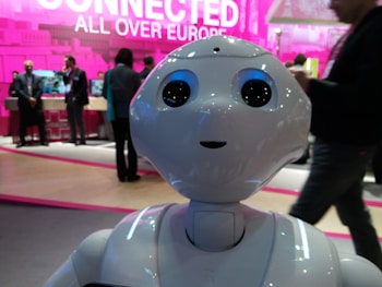 A humanoid robot with large, expressive eyes stands prominently, capturing attention in a tech event setting. People in business attire are visible in the background, engaged in conversation and examining displays. The scene is vibrant with a backdrop that features the phrase 'Connected All Over Europe' in bold pink lettering.