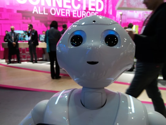 A humanoid robot with large, expressive eyes stands prominently, capturing attention in a tech event setting. People in business attire are visible in the background, engaged in conversation and examining displays. The scene is vibrant with a backdrop that features the phrase 'Connected All Over Europe' in bold pink lettering.