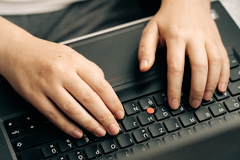 Two hands typing on a black laptop keyboard. The laptop has a red pointing stick in the middle of the keyboard.