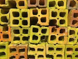 A stack of colorful cinder blocks featuring a mix of green, yellow, and orange hues. The blocks are arranged in a grid-like pattern, with their hollow sections prominently visible.