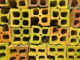 A stack of colorful cinder blocks featuring a mix of green, yellow, and orange hues. The blocks are arranged in a grid-like pattern, with their hollow sections prominently visible.
