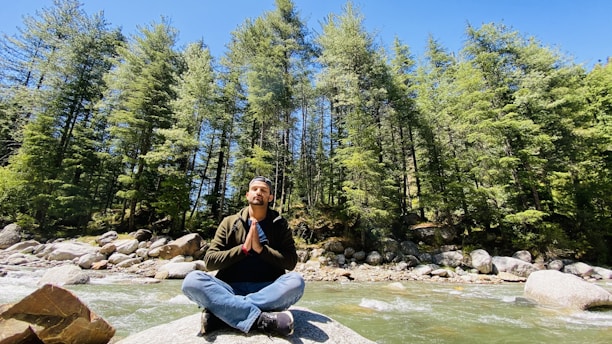 A person is sitting cross-legged on a rock near a flowing river, surrounded by dense evergreen trees. The individual appears to be meditating in a serene natural setting under a clear blue sky.
