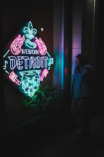 A vibrant neon sign in a window displays the words 'WeWork Detroit' alongside symbols like a fleur-de-lis, flamingo, and roaring tiger. The colors ripple in shades of pink, blue, and green, contrasting with the dark surroundings. A person stands to the right, wearing glasses and a hooded jacket, observing the sign amidst some foliage at the base.