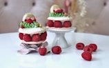 Two elegantly crafted desserts featuring pink macarons sandwiched with fresh raspberries are arranged on a white stand and tabletop. The macarons are adorned with green frosting, gold leaf, and additional decorative elements. A few loose raspberries are scattered around, adding a touch of freshness to the composition.