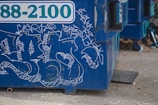 A blue dumpster features graffiti with various white, abstract designs and scribblings. The dumpster, with a partially visible telephone number, is situated on a concrete or gravel ground. The foreground shows some scattered debris and small objects.